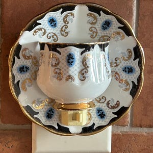 May include: A wall-mounted night light crafted from a white porcelain teacup and saucer featuring a black, gold, and light blue floral pattern. The piece includes a beige plastic plug-in base with an on-off switch centered at the bottom.