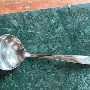 May include: A polished silver-tone metal ladle featuring a deep, rounded bowl and a slender handle. The handle is decorated with two small, engraved star-like motifs and subtle, flowing lines that extend toward the end of the utensil.