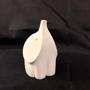 May include: A matte white ceramic figurine shaped like a stylized elephant. The object features a smooth, rounded body with a singular small circular protrusion representing an eye and a curved, tapered trunk extending upward from the head.
