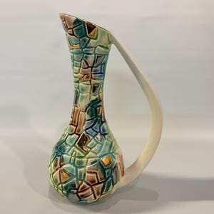 May include: A ceramic pitcher featuring a mosaic-like pattern of teal, brown, and cream segments with gold accents. The vessel has a tall, slender neck, a bulbous base, and a single, smooth, cream-colored handle extending from the rim to the body.