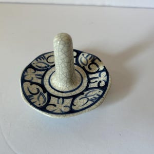 May include: A ceramic ring holder featuring a central vertical post and a circular base. The piece has a cream-colored glaze with a fine crackle finish and is decorated with dark blue hand-painted floral and leaf patterns around the base.