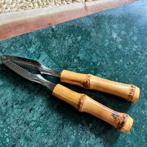 May include: A metal nutcracker featuring two hinged arms with serrated inner edges for gripping and light brown bamboo handles with natural textured nodes. The tool is designed with a silver-toned finish on the metal components and a smooth, polished appearance on the bamboo grips.