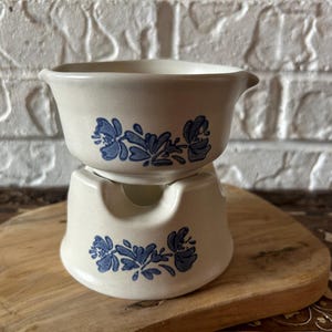 May include: A two-piece off-white ceramic fondue pot set featuring a bowl with a pouring spout and a matching base stand. Both pieces are decorated with a blue floral pattern consisting of stylized blossoms and leaves centered on the exterior.