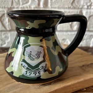 May include: A ceramic mug with a glossy camouflage pattern in shades of green, brown, and black. The front features a white emblem with crossed rifles and wheat stalks above a banner displaying the year 1775. It has a wide base and a black handle.