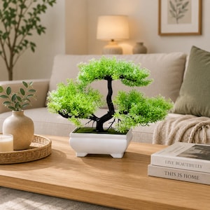 May include: A small artificial bonsai tree with vibrant green foliage and a dark, curved trunk. The tree is planted in a rectangular white ceramic pot with four small feet, featuring a moss-like base covering the soil surface.