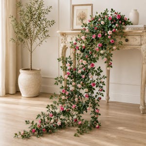 May include: A long, trailing artificial rose garland featuring numerous small, vibrant pink and soft cream-colored rose blossoms interspersed with lush green leaves. The vine cascades from the top of a wooden console table down to the floor.