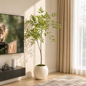May include: A tall artificial green tree with slender branches and pointed leaves, potted in a smooth, spherical white planter. The plant stands upright with a thin brown trunk, displaying a natural branching pattern and vibrant green foliage.