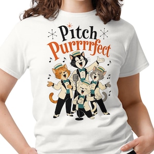 May include: A white short-sleeved t-shirt featuring a graphic of four cartoon cats dressed in formal suits and hats, singing into a microphone. The text "Pitch Purrrfect" is printed above the cats in black and orange, surrounded by musical notes.