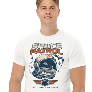 May include: A white t-shirt featuring a graphic print of an astronaut helmet with a moon landing reflection. The text reads SPACE PATROL at the top and DEEP SPACE OPERATIONS at the bottom, accompanied by a satellite, stars, and a stylized SP emblem.