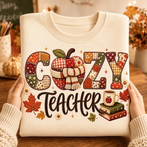May include: A cream-colored sweatshirt featuring the text COZY TEACHER in a patchwork-style font. The design includes a central apple wearing a scarf, a stack of two books, a mug with an apple graphic, and scattered autumn leaves and flowers.