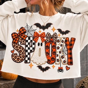 May include: A white long-sleeve sweatshirt featuring the word SPOOKY in large, decorative letters. Each letter has a unique pattern including leopard print, black and white checkers, a ghost, pumpkins, and orange plaid. Bows, bats, and webs accent the design.