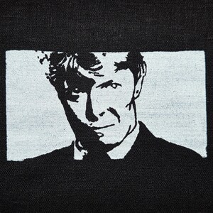 May include: A black and white textile print featuring a high-contrast, stencil-style portrait of a person with short, textured hair. The image is framed within a rectangular white border set against a solid black fabric background.