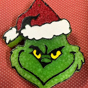 May include: A green, textured character-shaped ornament wearing a red glittery Santa hat with a white brim. The character has black outlines, a wide grin, and narrow yellow eyes. The surface of the green portion is bumpy and translucent in appearance.