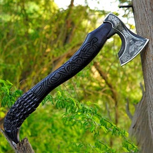 Può includere: Un'ascia a mano con un manico in legno scuro intagliato che presenta intricati motivi a nodo e a scaglie. La testa dell'ascia in metallo mostra dettagliate incisioni color argento di nodi celtici e un motivo centrale a uccello.