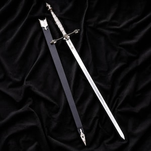 May include: A silver-toned metal sword with a textured grip and a cross-shaped guard, positioned alongside a long, slender black scabbard. The scabbard features a metallic silver-coloured throat and a pointed metal tip at the bottom end.