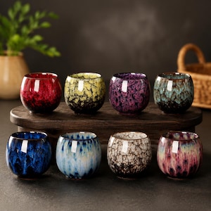 May include: A set of eight small, round ceramic tea cups featuring a variety of vibrant, multi-coloured crackle glaze finishes. The cups are arranged in two rows on a dark wooden stand, displaying colours including red, yellow, purple, teal, blue, and white.