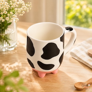 May include: A ceramic mug featuring a black and white cow print pattern, a smooth white interior, and three small, rounded light pink feet at the base. The handle is white with a black spot, and the mug has a glossy finish throughout.