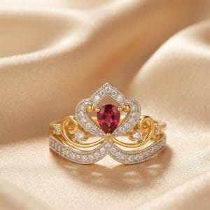 May include: A gold ring featuring a crown-shaped design adorned with small clear stones and a single pear-shaped red gemstone at the center. The band and the crown structure are encrusted with pavé-set clear stones, creating a detailed, layered look.
