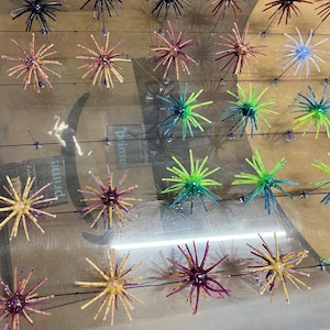 May include: A collection of multi-colored silicone fishing skirt lures arranged in rows on a clear plastic sheet. The lures feature thin, radiating strands in various colors including black, brown, purple, yellow, blue, and green, with some speckled patterns.