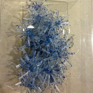 May include: A collection of clear, star-shaped plastic ornaments with blue glitter accents, contained within a transparent rectangular plastic box. Each piece features multiple radiating points extending from a central core, creating a spiky appearance.