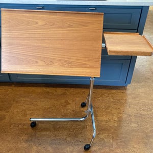 May include: A mobile adjustable desk featuring a large rectangular wood-grain surface and a smaller attached side tray. The unit is supported by a chrome-plated metal frame with a curved base and four black caster wheels for easy movement.