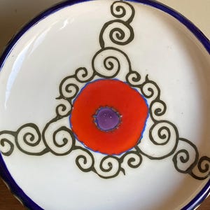 May include: A decorative ceramic plate with a white base, featuring a central red circle with a purple center, outlined in blue. Black swirl designs surround the red circle. The rim is a deep blue.