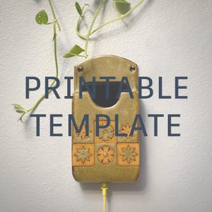 May include: A wall-mounted ceramic phone holder with a mottled olive green glaze and a grid of six square tiles featuring embossed flower patterns. The holder has a central cutout for a phone and a bottom opening for a charging cable to pass through.