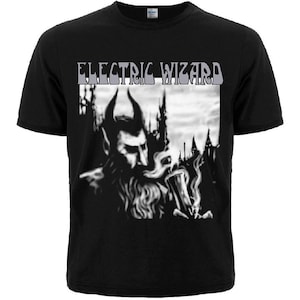 May include: A black short-sleeved t-shirt featuring a graphic print with the text ELECTRIC WIZARD in a stylised font. The image depicts a dark, horned figure with a beard, surrounded by smoke and silhouettes of pointed structures in the background.