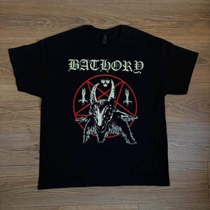 Może przedstawiać: Czarny t-shirt z krótkim rękawem z nadrukiem graficznym na klatce piersiowej. Wzór zawiera słowo BATHORY wypisane białą gotycką czcionką nad czerwonym pentagramem, wewnątrz którego znajduje się ilustracja białej głowy kozła z czerwonymi oczami i dwoma mieczami.