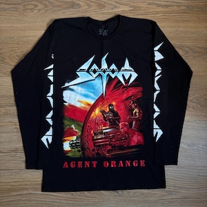 May include: A black long-sleeve t-shirt featuring a graphic print of the band Sodom. The design includes the band name in white, a colourful war-themed illustration with soldiers and artillery, and the text AGENT ORANGE in red at the bottom.