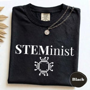Puede incluir: Una camiseta negra con el texto blanco STEMinist sobre un gráfico blanco de un chip de computadora. Un collar de cadena plateada con un colgante redondo descansa sobre el cuello. La camiseta tiene una etiqueta de marca Comfort Colors.