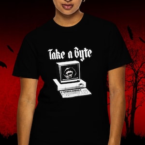 May include: A black short-sleeved t-shirt featuring a white graphic of a vintage computer with a pair of vampire fangs displayed on the screen. Above the computer, the text "Take a Byte" is printed in a bold, gothic-style white font.