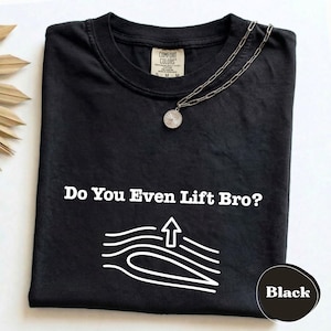 May include: A black t-shirt featuring the white text Do You Even Lift Bro? above a white line graphic of an airfoil with an upward arrow. A silver-toned chain necklace with a round pendant rests on the neckline of the shirt.