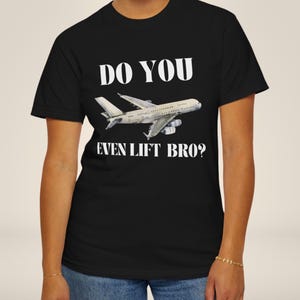 Pode incluir: Uma t-shirt preta de manga curta com um gráfico de um avião de passageiros branco ao centro. Acima do avião, o texto diz DO YOU, e abaixo do avião, o texto diz EVEN LIFT BRO? em letras maiúsculas brancas e em negrito.