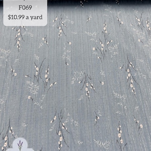 Peut inclure: Un tissu bleu poussiéreux avec un motif répétitif de petites fleurs blanches et de fines tiges sombres. Le matériau présente une subtile texture côtelée verticale. Le texte sur l'image indique : 100% poly, dusty blue, F069, $10.99 a yard, Farmgirl DRESSES.