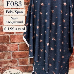 Puede incluir: Una tela azul marino con un pequeño estampado floral de flores rosas y blancas. El texto en la imagen dice F083, Poly/Span, Navy background, $11.99 a yard y Farmgirl Dresses. La tela tiene una apariencia texturizada y bordes blancos.