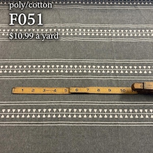 May include: A grey poly/cotton fabric featuring horizontal white stripes and a repeating white triangle pattern. A yellow measuring tape is placed across the fabric, showing a length of 28 cm. The text poly/cotton F051 is visible.