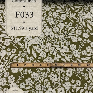 May include: Olive green fabric featuring a white floral and leaf pattern. A wooden ruler is placed horizontally across the fabric, showing a measurement of 10 inches. Text on the fabric reads Cotton/linen, F033, and FARMGIRL DRESSES LLC.