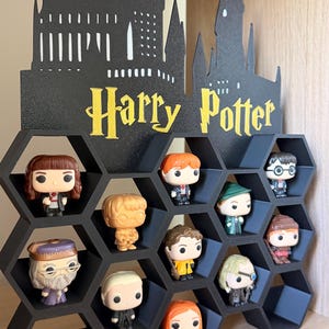 May include: A black honeycomb-shaped display shelf featuring a silhouette of Hogwarts castle and the yellow text Harry Potter. The shelf contains eleven small vinyl character figurines with large heads and eyes arranged in the hexagonal compartments.