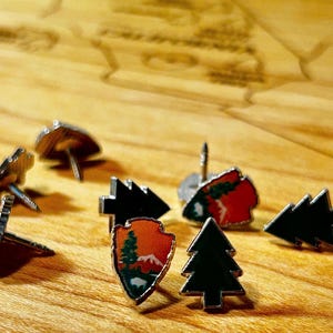 Puede incluir: Una colección de pines metálicos con formas de punta de flecha con diseños de paisajes montañosos en naranja y rojo, y formas de árboles de hoja perenne en negro sólido. Cada pin tiene un borde de metal plateado y un poste metálico.