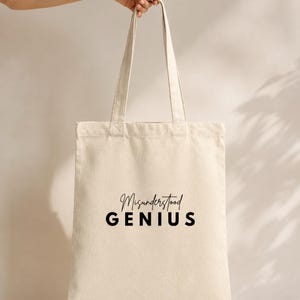 Puede incluir: Un bolso de tela de color crema con dos correas para el hombro. La parte frontal del bolso presenta el texto Misunderstood GENIUS impreso en negro, con la palabra Misunderstood en fuente cursiva y GENIUS en una fuente negrita sin remates.