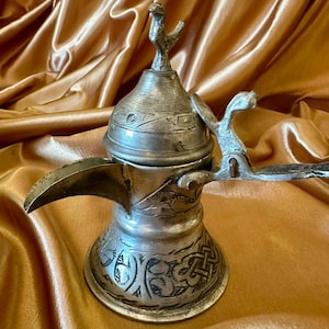 May include: A silver-toned metal coffee pot featuring a long, curved spout, a hinged lid topped with a small bird-shaped finial, and an attached handle. The body and lid are decorated with intricate, dark-etched geometric and floral patterns.