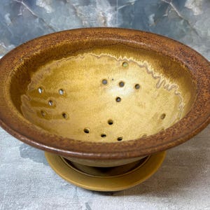 May include: A round ceramic berry bowl with a wide, speckled brown rim and a smooth, golden-yellow interior. The bowl features multiple drainage holes arranged in a circular pattern across the base and sits on a matching circular yellow saucer.