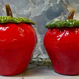 May include: Two ceramic strawberry-shaped canisters with glossy red bodies and textured green leaf lids. Each container features a small brown stem protruding from the center of the lid. The canisters have a rounded, bulbous form and a smooth finish.