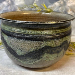 May include: A round ceramic bowl with a wide rim, featuring a textured exterior with a mottled glaze in shades of sage green, charcoal black, and earthy beige. The interior of the bowl has a smooth, light brown finish with a visible rim edge.