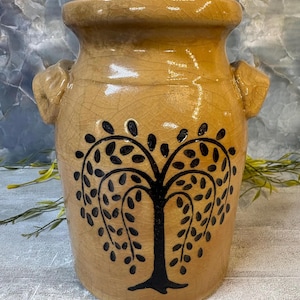 May include: A tan ceramic crock with a glossy crackle glaze finish, featuring two side handles and a black painted tree design on the front. The vessel has a wide, rounded rim and a cylindrical body, showcasing a distinct network of fine lines.