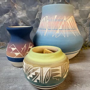 May include: Three ceramic vessels with matte finishes and etched geometric patterns. The largest is blue and pink, the medium is blue and brown, and the smallest is green and brown. Each piece features a white base and intricate carvings.