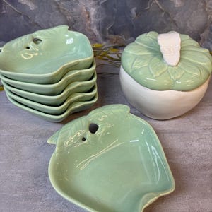 May include: A set of light green ceramic dishes shaped like apples, including a stack of five small plates and one larger lidded container with a white stem handle. The dishes feature a glossy finish and a molded leaf pattern on the lid.