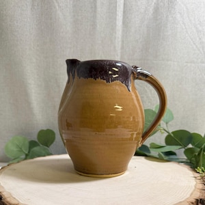 May include: A ceramic pitcher featuring a smooth, light tan body and a dark brown glazed rim that drips down the upper portion. The vessel has a rounded, bulbous shape with a single curved handle on the right side and a small pouring spout.