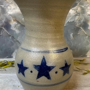 May include: A stoneware vase with a speckled light gray finish and a flared rim. The body features a horizontal blue band near the top and bottom, with three dark blue painted stars and small dots encircling the center of the rounded vessel.